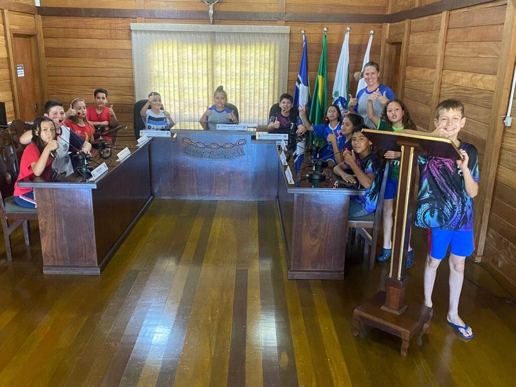 Nesta foto os alunos estão descontraídos, ocupando as cadeiras dos vereadores e o púlpito onde são realizadas as falas durante as sessões. Ao fundo, as bandeiras do Brasil, do Estado do Paraná, do Município de Porto Vitória e do Poder Legislativo compõem o cenário do plenário da Câmara