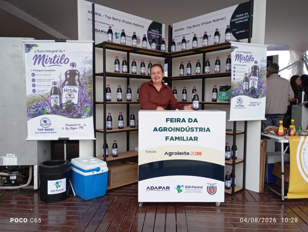 A Vereadora Elza está posicionada atrás de um banner, no qual está escrito: Feira da agroindústria familiar, ao fundo, produtos coloniais estão espostos em prateleiras.